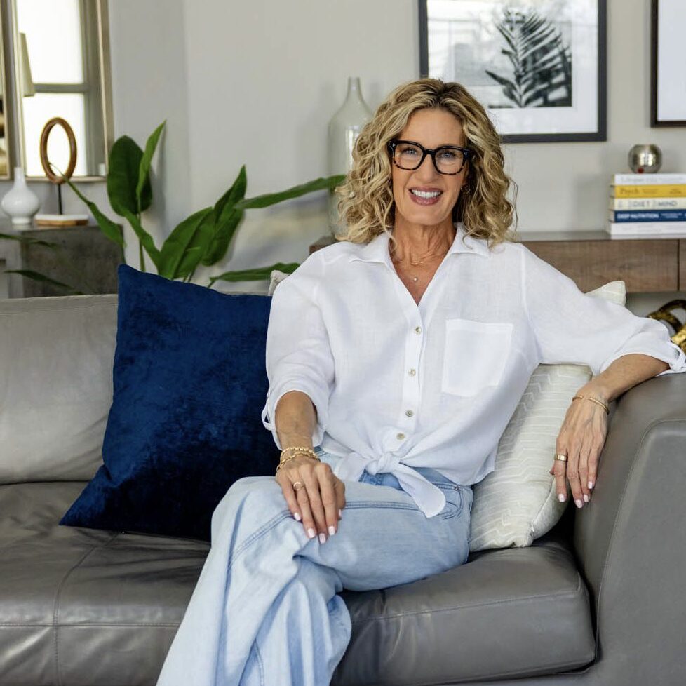 GoDaddy_-5418 2 a midlife woman sitting on the sofa in her peaceful home embodying a healthy and happy outlook while wearing a white linen tie-top and light-colored jeans.